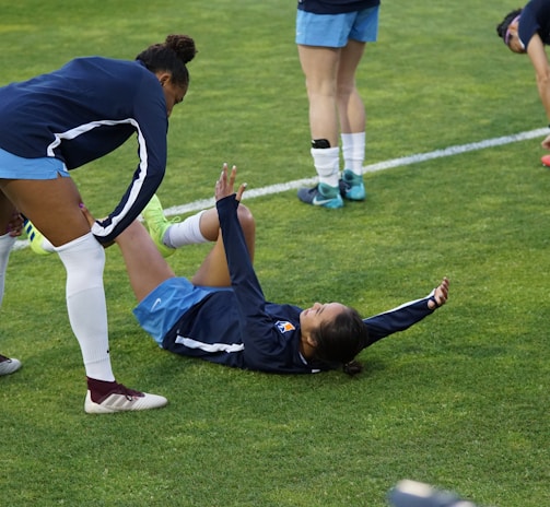 A group of athletes in sportswear are on a green soccer field. Two individuals are interacting closely; one helps the other who is lying on the grass. Nearby, other athletes are stretching.