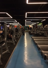 A spacious gym with multiple rows of treadmills and weightlifting benches on either side. The lighting features geometric patterns on the ceiling, creating a modern and organized ambiance. People are actively using the equipment, engaging in fitness activities.