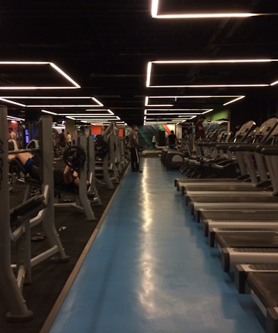 A spacious gym with multiple rows of treadmills and weightlifting benches on either side. The lighting features geometric patterns on the ceiling, creating a modern and organized ambiance. People are actively using the equipment, engaging in fitness activities.