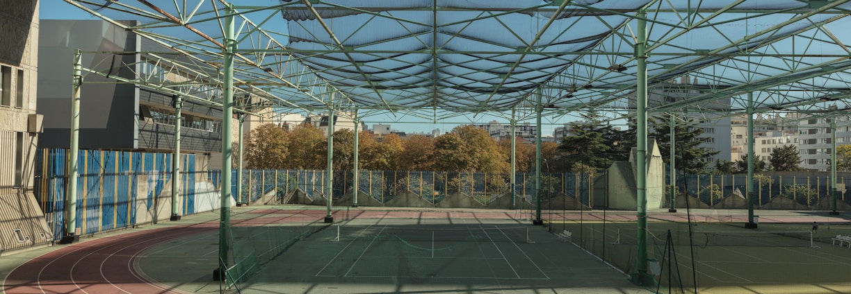 A spacious indoor sports facility with a tennis court and running tracks. The court is surrounded by high nets and tall, green metal structures supporting a roof. The area is well-lit with natural light filtering in through a transparent ceiling. In the background, there are trees with autumn foliage and several residential buildings.