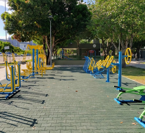 An outdoor fitness area featuring a variety of exercise equipment, including stationary bikes and other workout stations. The equipment is primarily blue and yellow, arranged neatly on a paved surface. Surrounding the area are trees providing shade, with a few parked cars and buildings visible in the background.