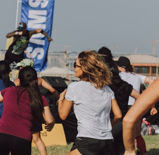 A group of people appears to be participating in an outdoor fitness class or dance session. They are dressed in athletic clothing, and a blue banner can be seen in the background, along with a stage. The setting suggests a lively and energetic atmosphere.