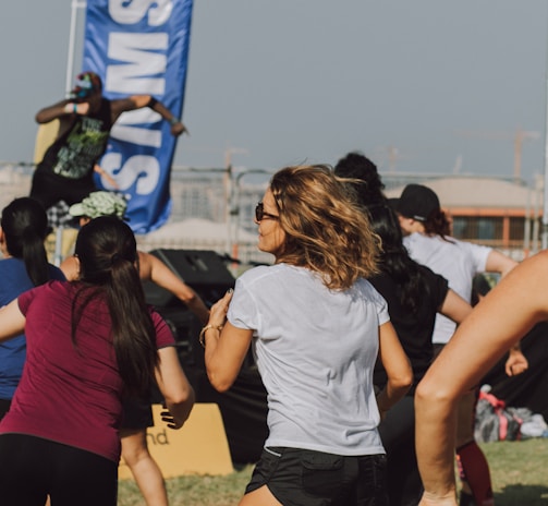 A group of people appears to be participating in an outdoor fitness class or dance session. They are dressed in athletic clothing, and a blue banner can be seen in the background, along with a stage. The setting suggests a lively and energetic atmosphere.