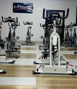 A room filled with stationary exercise bikes arranged neatly in rows. The space has a wooden and gray tiled floor, and a sign with the text 'i-Fitness' is displayed on the wall. The bikes are primarily white with black handlebars and seats.