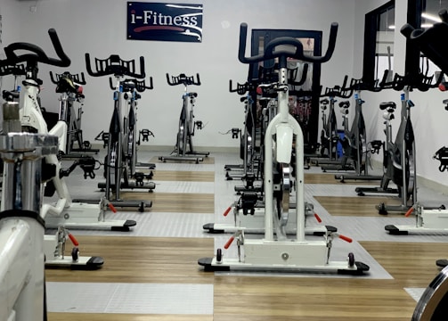 A room filled with stationary exercise bikes arranged neatly in rows. The space has a wooden and gray tiled floor, and a sign with the text 'i-Fitness' is displayed on the wall. The bikes are primarily white with black handlebars and seats.