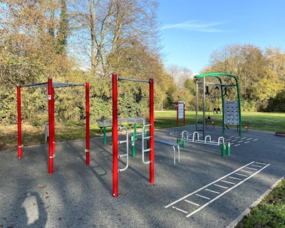 An outdoor fitness park surrounded by trees features various exercise stations with equipment like bars and rings. The area is bordered by lush green grass and there are tall trees with autumn-colored leaves in the background. The sky is clear and blue, creating a serene environment ideal for workouts.