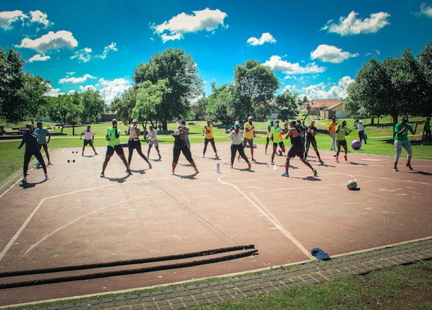 A group of people are exercising outdoors on a concrete court, surrounded by green spaces and trees. They are standing in a formation, appearing to engage in an organized fitness class. Each participant wears athletic clothing, and several carry exercise equipment such as dumbbells and ropes. The sky is bright blue with scattered fluffy clouds, creating a vibrant and energetic atmosphere.