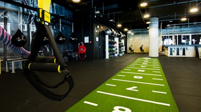 A modern gym space featuring a green track laid out on the floor with numbered lanes. TRX suspension trainers hang prominently in the foreground. The gym is equipped with various exercise equipment, including kettlebells and punching bags. In the background, there is a FIT sign near a reception desk under a low-ceilinged industrial design.