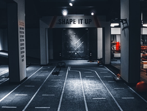 A dimly lit indoor gym with a central pathway marked with lane lines. The ceiling features multiple lights, and there are exercise equipment and motivational signage, including one that reads 'SHAPE IT UP'. The ambiance suggests a focus area for fitness activities.