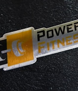 A metal plaque with the text 'POWER FITNESS' is set against a textured black background. The design includes a stylized dumbbell icon adjacent to the text, with a modern and sleek appearance.