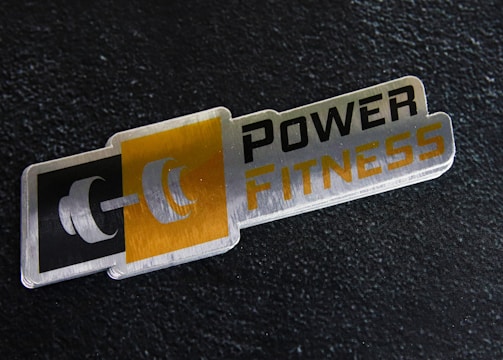 A metal plaque with the text 'POWER FITNESS' is set against a textured black background. The design includes a stylized dumbbell icon adjacent to the text, with a modern and sleek appearance.