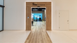 A modern fitness center entrance with wooden framing and the sign 'PRIME FITNESS' above the doorway. The interior features a spacious gym area with workout equipment visible and a motivational banner on the back wall. The flooring transitions from glossy tiles to a wood finish leading into the facility.