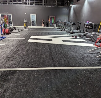 A spacious gym with a black and white color scheme featuring various fitness equipment. On the left, numerous weight plates and bars are organized on racks. In the center, a large open workout area is visible with a textured floor and white directional markings. To the right, there are treadmills, stationary bikes, and other workout machines along with large exercise balls. Exercise bands and weightlifting belts are hanging on hooks at the back wall. A single person is seen near a door, adding a human element to the scene.