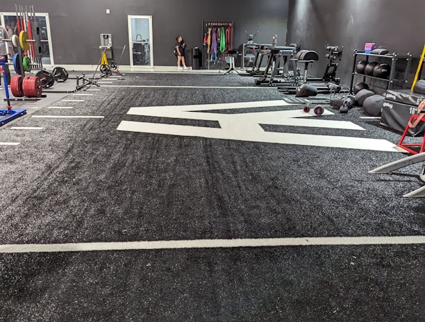 A spacious gym with a black and white color scheme featuring various fitness equipment. On the left, numerous weight plates and bars are organized on racks. In the center, a large open workout area is visible with a textured floor and white directional markings. To the right, there are treadmills, stationary bikes, and other workout machines along with large exercise balls. Exercise bands and weightlifting belts are hanging on hooks at the back wall. A single person is seen near a door, adding a human element to the scene.