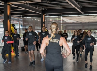 A group of individuals in athletic wear participate in a fitness class inside a spacious, well-lit room. The instructor, facing away, leads the group with energy and focus. The participants appear engaged and attentive, following along with the instructor's movements.