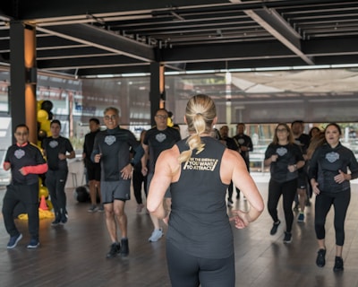 A group of individuals in athletic wear participate in a fitness class inside a spacious, well-lit room. The instructor, facing away, leads the group with energy and focus. The participants appear engaged and attentive, following along with the instructor's movements.