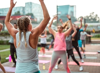 A group of people is participating in an outdoor fitness class on a paved area with yoga mats laid out. In the foreground, a person with braided hair, wearing a sleeveless top and leggings, raises their arms. Other participants are dressed in sportswear, energetically following the movements of the instructor.