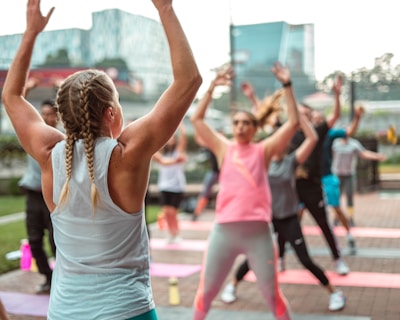 A group of people is participating in an outdoor fitness class on a paved area with yoga mats laid out. In the foreground, a person with braided hair, wearing a sleeveless top and leggings, raises their arms. Other participants are dressed in sportswear, energetically following the movements of the instructor.