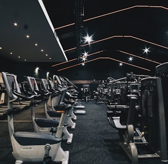 A spacious gym filled with various exercise equipment. Treadmills are aligned in rows on one side, while weight machines and other fitness devices are organized on the other side. The ceiling features modern, angular lines with bright overhead lights, adding a sleek and contemporary feel.