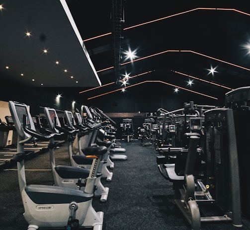 A spacious gym filled with various exercise equipment. Treadmills are aligned in rows on one side, while weight machines and other fitness devices are organized on the other side. The ceiling features modern, angular lines with bright overhead lights, adding a sleek and contemporary feel.