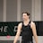 A person wearing a sleeveless black sports top is standing indoors in a gymnasium. The background includes a gray wall with green and white lettering partially visible.
