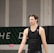 A person wearing a sleeveless black sports top is standing indoors in a gymnasium. The background includes a gray wall with green and white lettering partially visible.