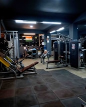 A dimly lit gym with various exercise machines, including leg presses and cable equipment. The floor is padded with dark tiles. In the background, two people are sitting at a desk with a computer. An Indian flag is visible on the wall, adding a touch of decoration.