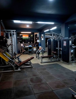 A dimly lit gym with various exercise machines, including leg presses and cable equipment. The floor is padded with dark tiles. In the background, two people are sitting at a desk with a computer. An Indian flag is visible on the wall, adding a touch of decoration.