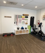 A fitness room featuring a variety of workout equipment neatly organized. The wall has a pegboard displaying exercise bands, foam rollers, and ropes. Below the board, there are kettlebells and dumbbells on shelves. An exercise bike is positioned to the right, with a step platform on the left.