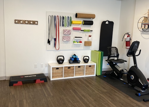 A fitness room featuring a variety of workout equipment neatly organized. The wall has a pegboard displaying exercise bands, foam rollers, and ropes. Below the board, there are kettlebells and dumbbells on shelves. An exercise bike is positioned to the right, with a step platform on the left.