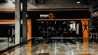 A fitness center entrance with the branding 'smart fit' illuminated above the doors. The interior has a modern design, featuring dark glass and bright orange accents. A reflective marble floor extends in front, capturing reflections from the surroundings. There are two individuals standing and walking towards the left.