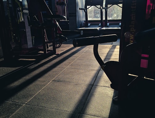 A dimly lit gym space with various fitness equipment including treadmills and weight machines. The sunlight creates long shadows on the floor, adding contrast to the scene.